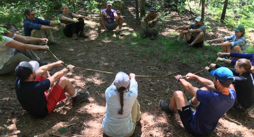 A group of people sit in a circle outdoors. They are all holding a rope and are participating in a team building exercise. 